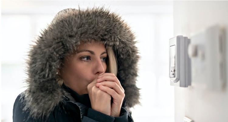 Woman wearing a parka, shivering in front of a broken thermostat - indicating a broken furnace